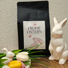 Schwarze Kaffeeverpackung der „Villa Rica Edition – Frohe Ostern“ mit Osteretikett (Hase und Schmetterlinge), dekoriert mit weißen Porzellan-Hasen, bunten Tulpen und gerösteten Kaffeebohnen auf einem Tisch – frühlingshafte Osterdekoration.