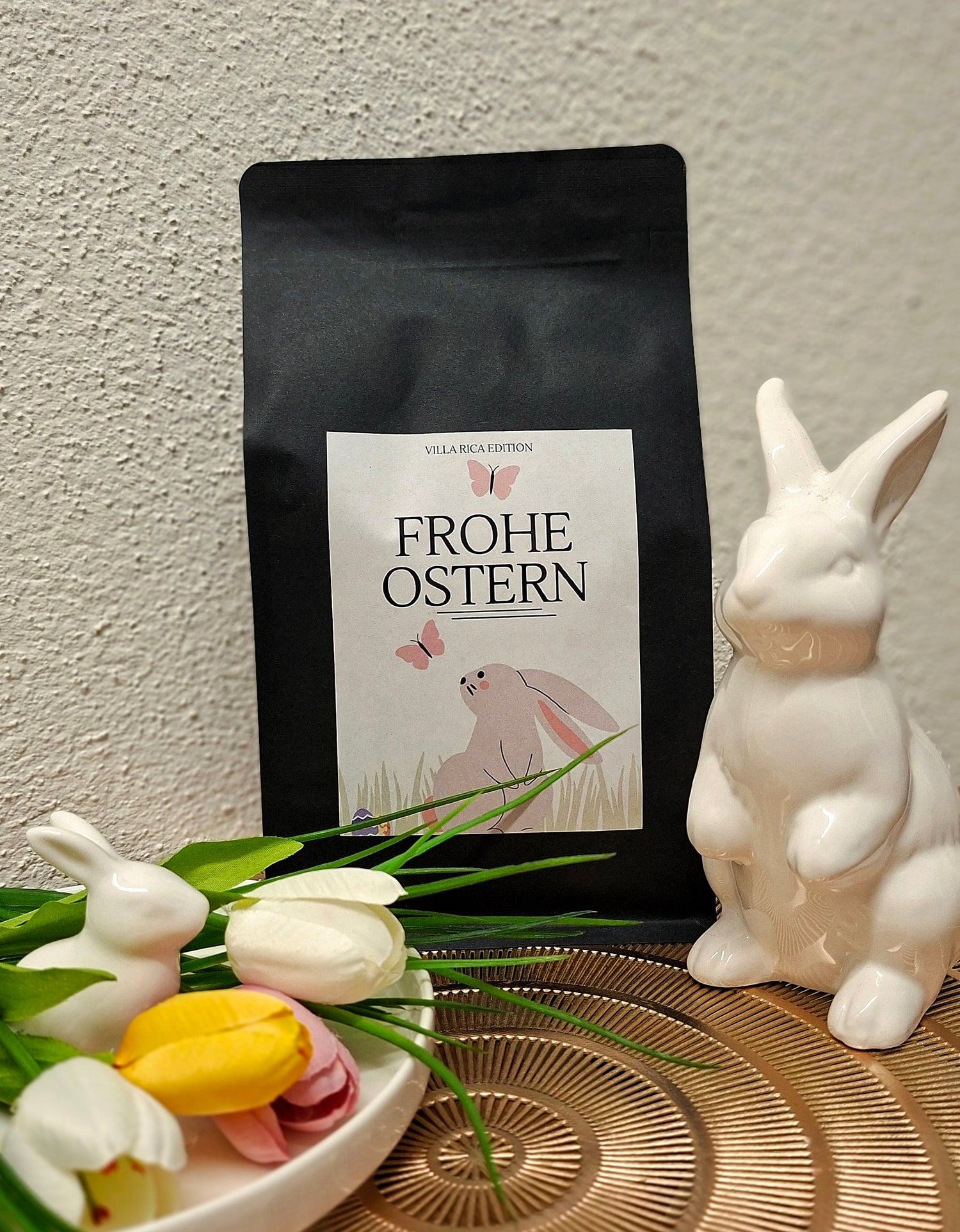 Schwarze Kaffeeverpackung der „Villa Rica Edition – Frohe Ostern“ mit Osteretikett (Hase und Schmetterlinge), dekoriert mit weißen Porzellan-Hasen, bunten Tulpen und gerösteten Kaffeebohnen auf einem Tisch – frühlingshafte Osterdekoration.
